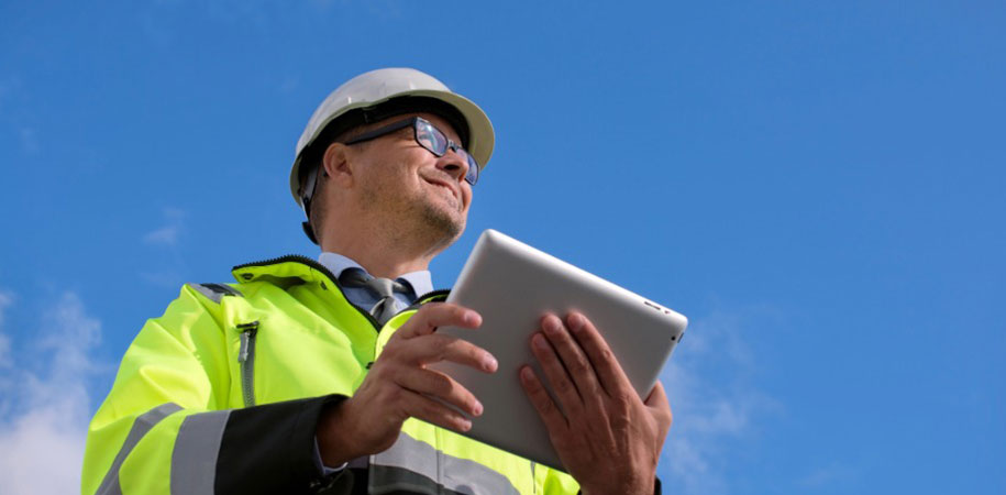 Construction inspector in a green safety coat and hard hat holding a tablet smiling and looking into the distance.