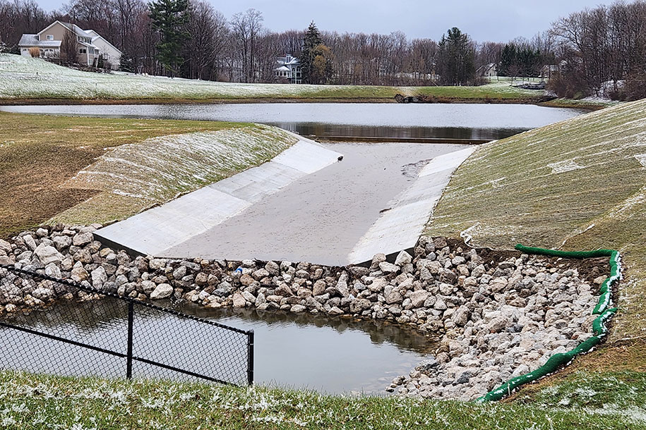 A lake in the background with water running over a low-sloped concrete spillway into a pool surrounded by rocks.