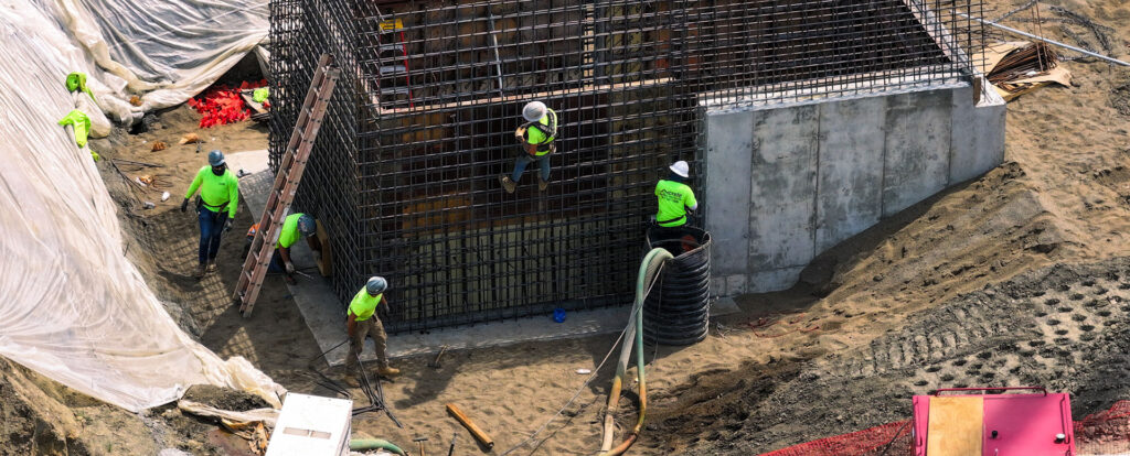 Construction workers in high-visibility gear assemble and secure a large reinforced steel structure beside a poured concrete wall, working on ladders and within the excavation area surrounded by sand, equipment, and protective tarps.