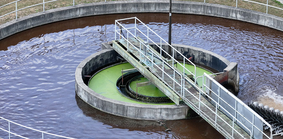 A circular wastewater treatment basin with a central mechanical arm and walkway, showing brown aerated water surrounding an inner channel coated with green algae or biofilm.