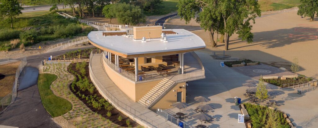 Aerial view of a modern, curved-roof pavilion at Edgewater Park in Cleveland, featuring an open-air upper deck, shaded outdoor seating with umbrellas, landscaped greenery, and surrounding walking paths near the lakefront.