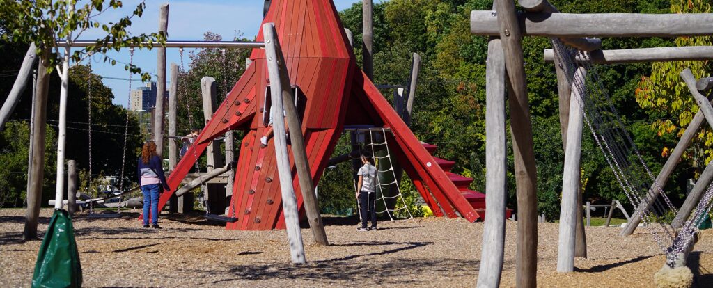 Children playing on a modern playground with a large red climbing structure, surrounded by wooden posts, rope features, and trees on a sunny day.