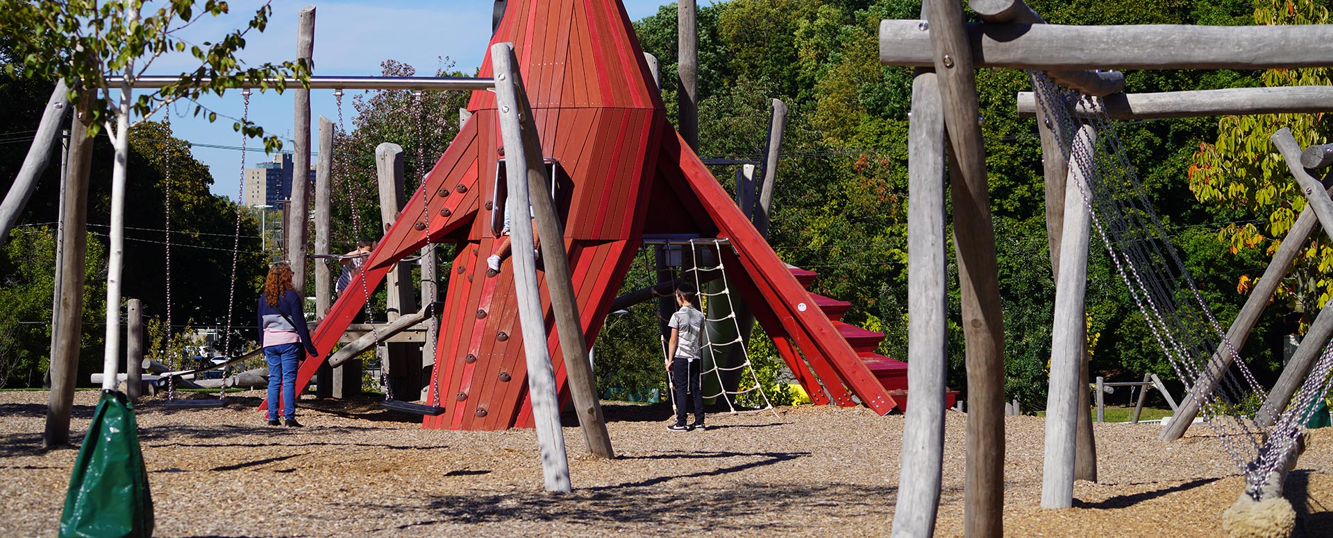 Children playing on a modern playground with a large red climbing structure, surrounded by wooden posts, rope features, and trees on a sunny day.