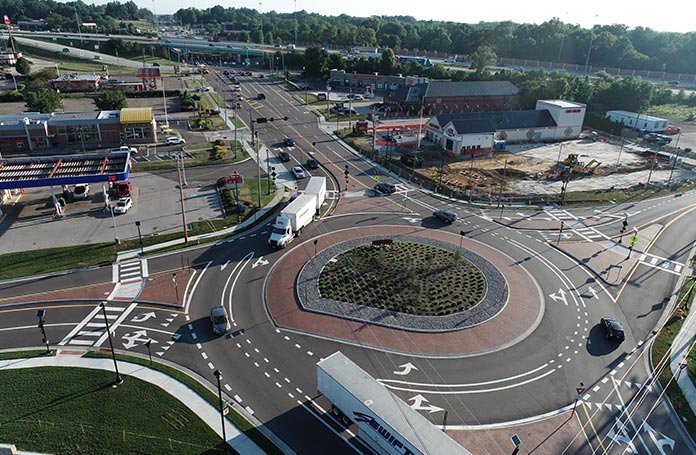 Aerial view of a landscaped roundabout with multiple entry roads, pedestrian crosswalks, directional lane markings, and a planted central island.