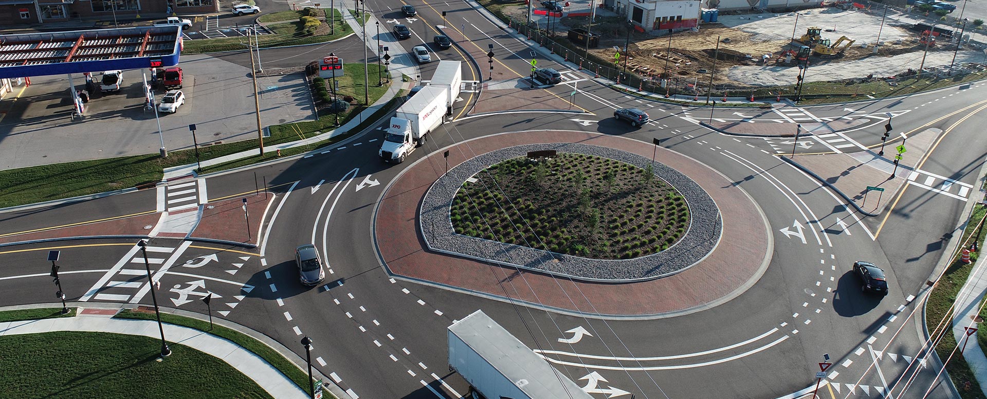 Aerial view of a landscaped roundabout with multiple entry roads, pedestrian crosswalks, directional lane markings, and a planted central island.